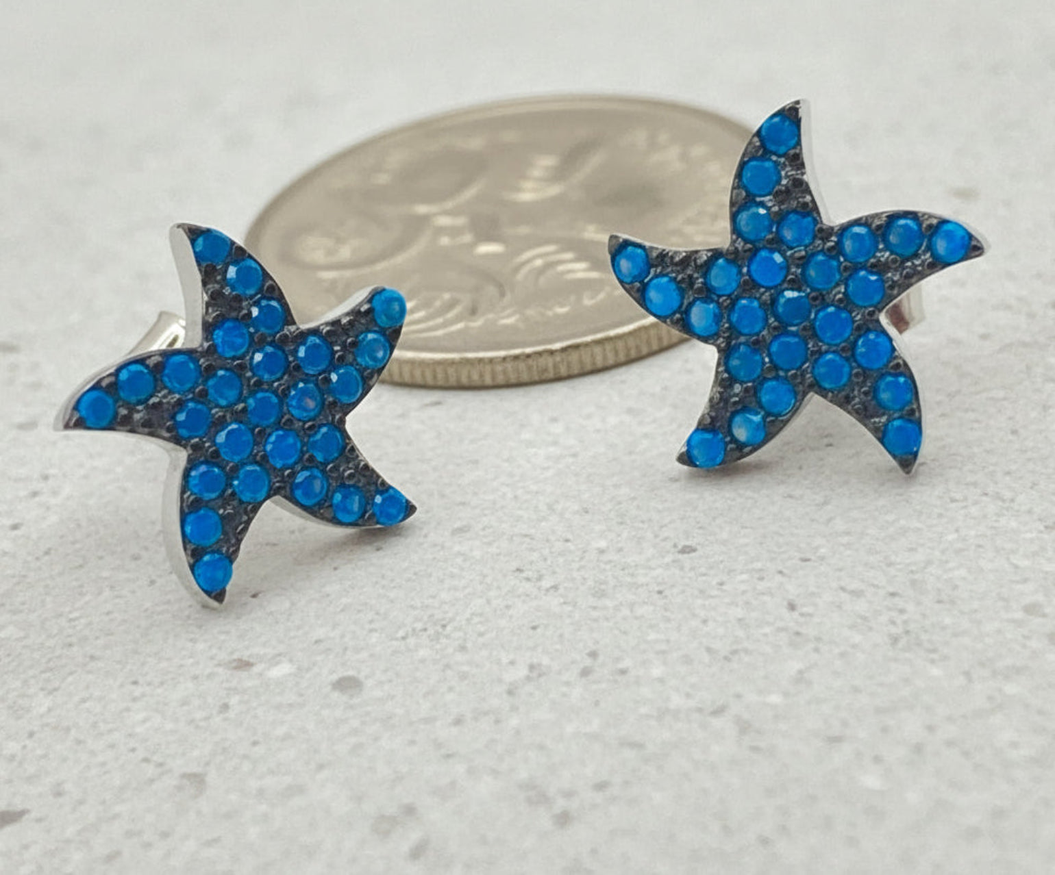 A pair of silver starfish stud earrings paved with bright blue rhinestones, shown next to a silver coin for scale on a flat surface.