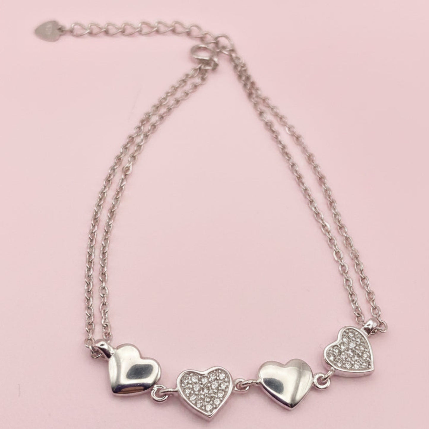 A silver double-strand chain bracelet featuring three heart charms, two of which are paved with small clear stones, displayed on a white background.