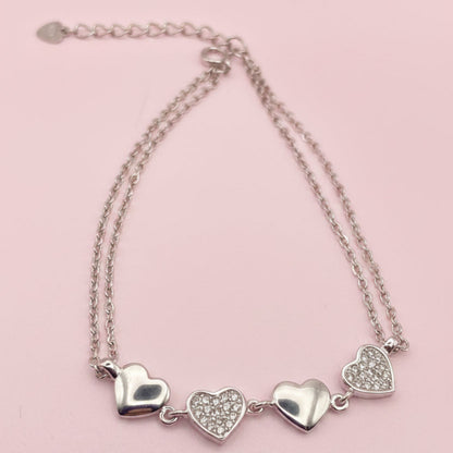 A silver double-strand chain bracelet featuring three heart charms, two of which are paved with small clear stones, displayed on a white background.