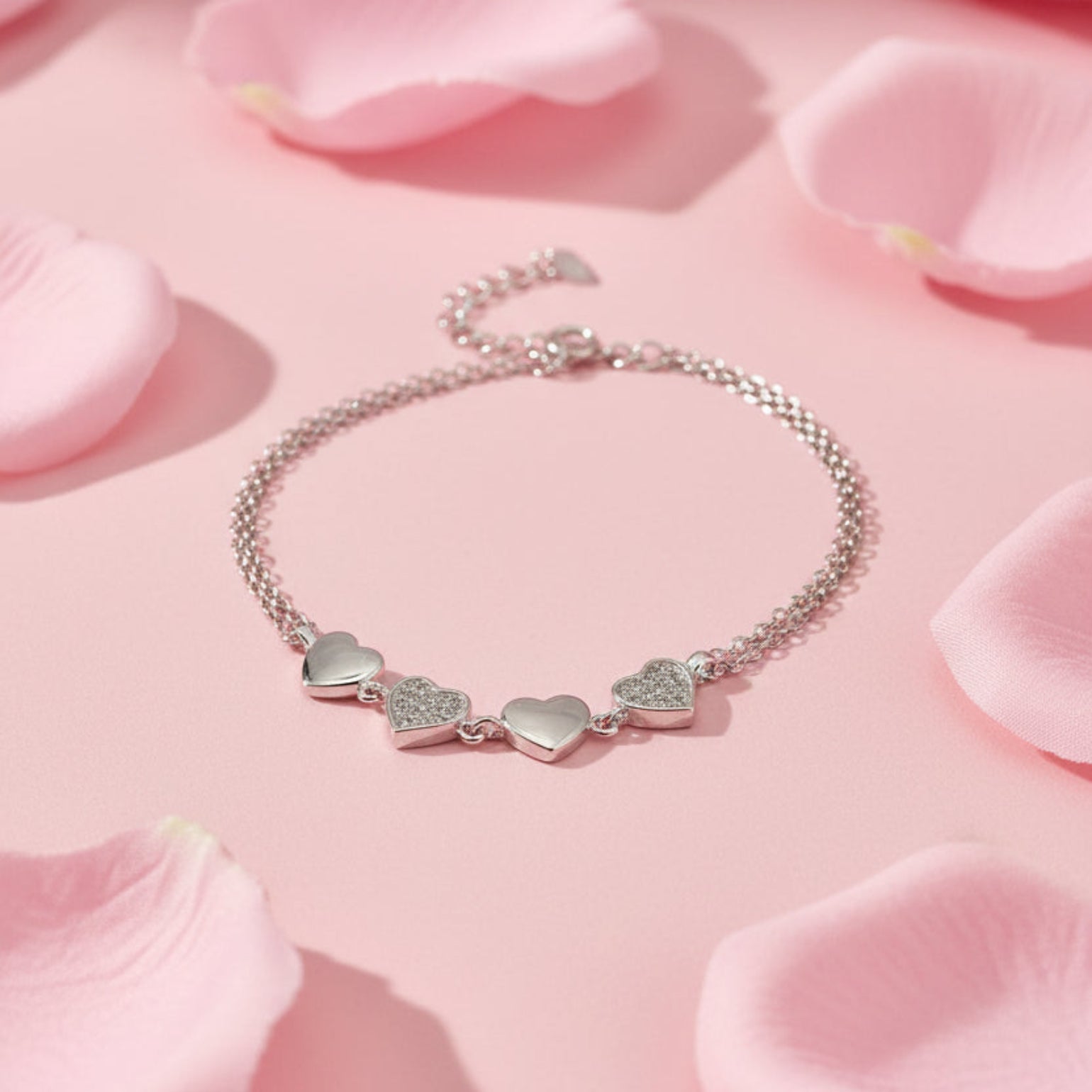 A silver chain bracelet with three heart charms, including two small heart charms covered in clear pavé stones, displayed on a white surface.