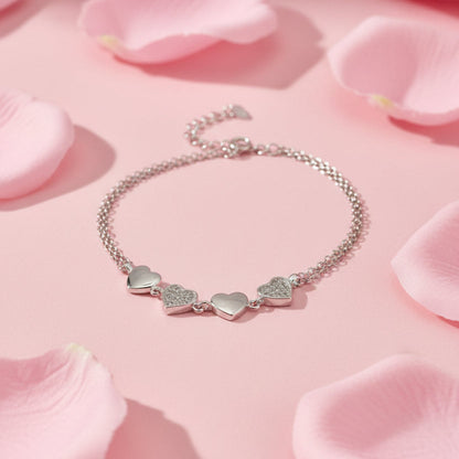 A silver chain bracelet with three heart charms, including two small heart charms covered in clear pavé stones, displayed on a white surface.