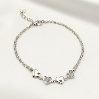 A silver chain bracelet with three dangling heart charms, displayed on a white satin fabric background.