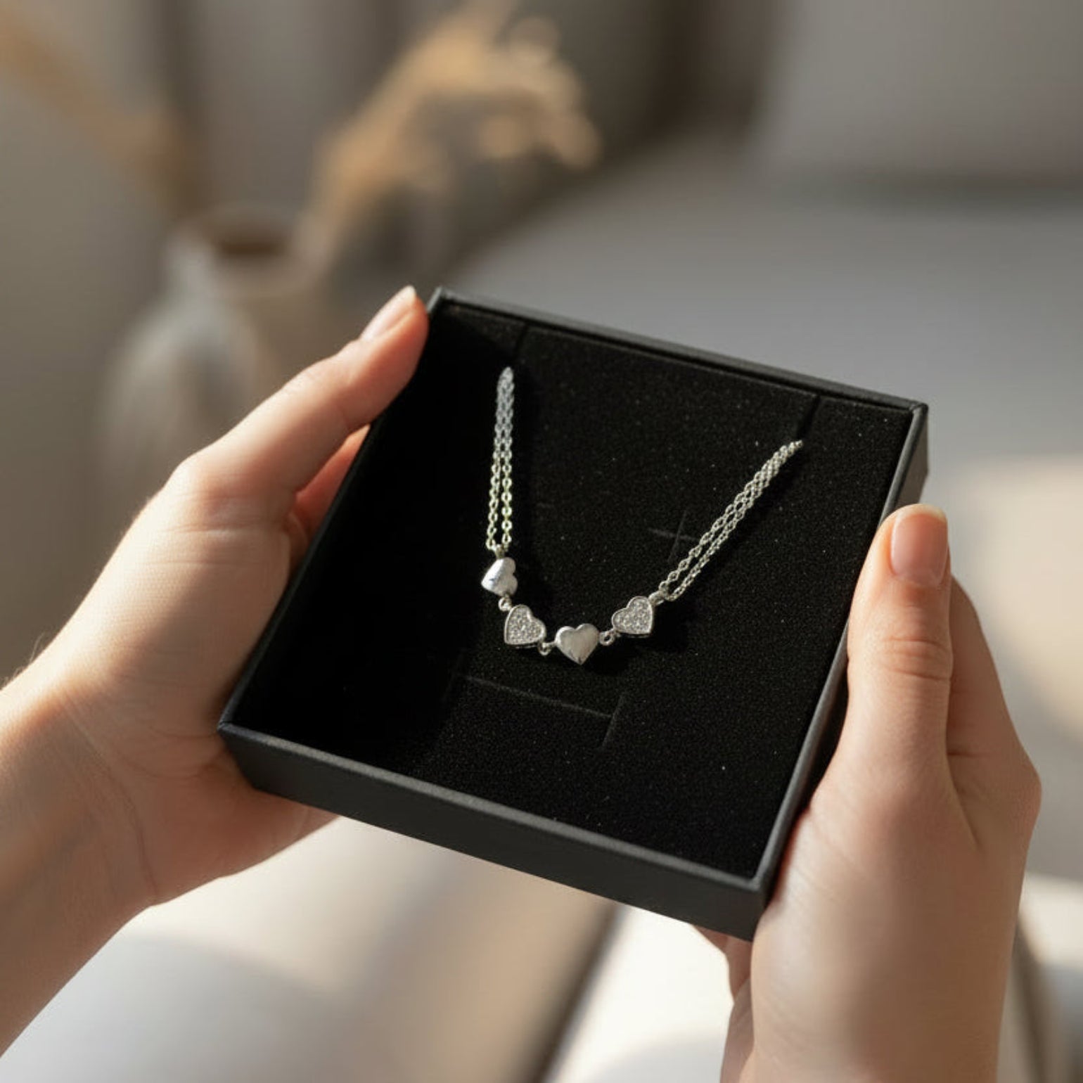 A silver heart-link necklace displayed in an open black jewelry box, held by two hands in a softly lit indoor setting.
