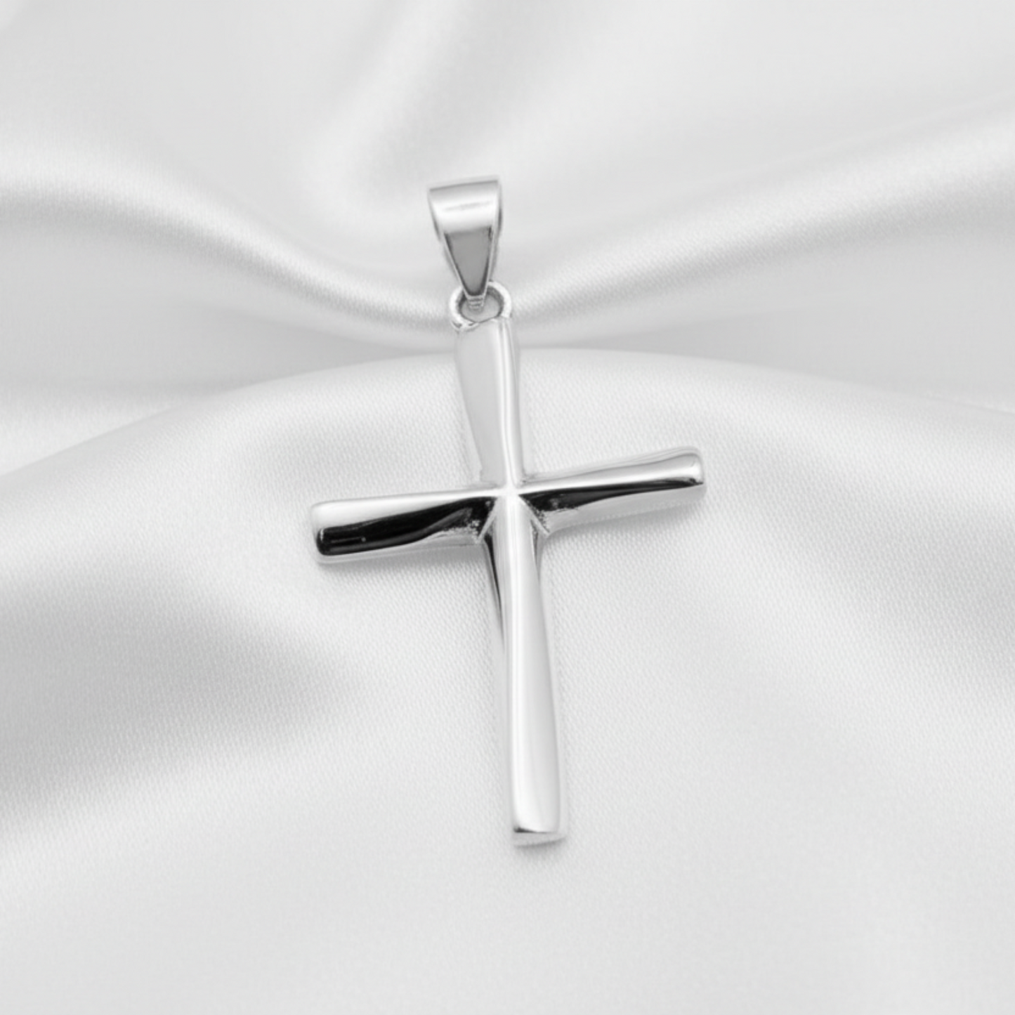 A polished silver cross pendant with a smooth, reflective finish, resting on a draped white satin fabric background.