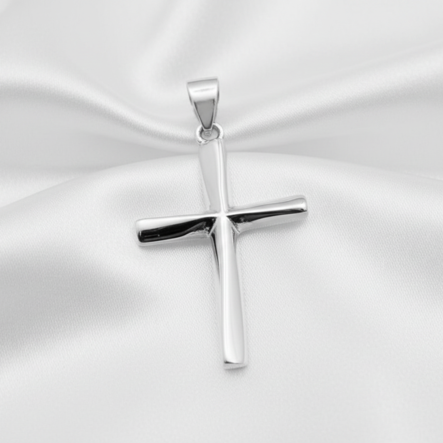 A polished silver cross pendant with a smooth, reflective finish, resting on a draped white satin fabric background.