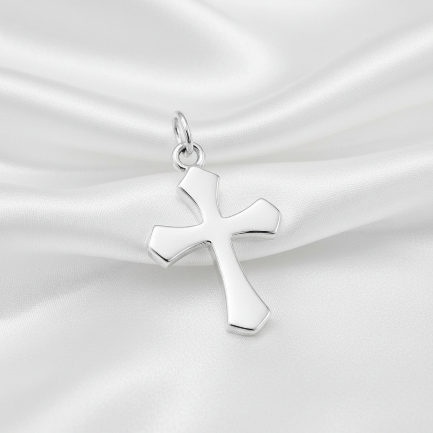A polished silver cross pendant with a smooth, reflective finish, resting on draped white satin fabric.