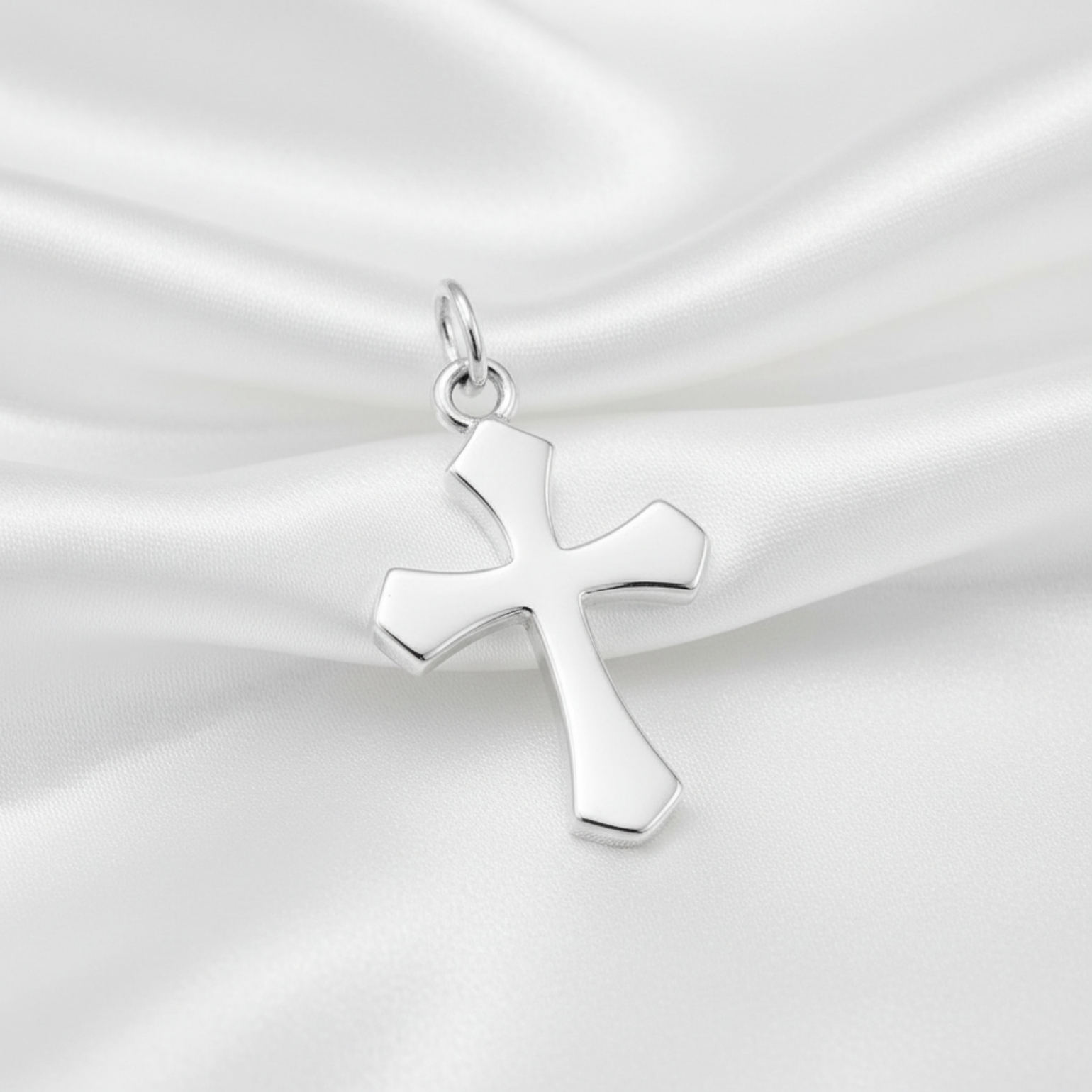 A polished silver cross pendant with a smooth, reflective finish, resting on draped white satin fabric.