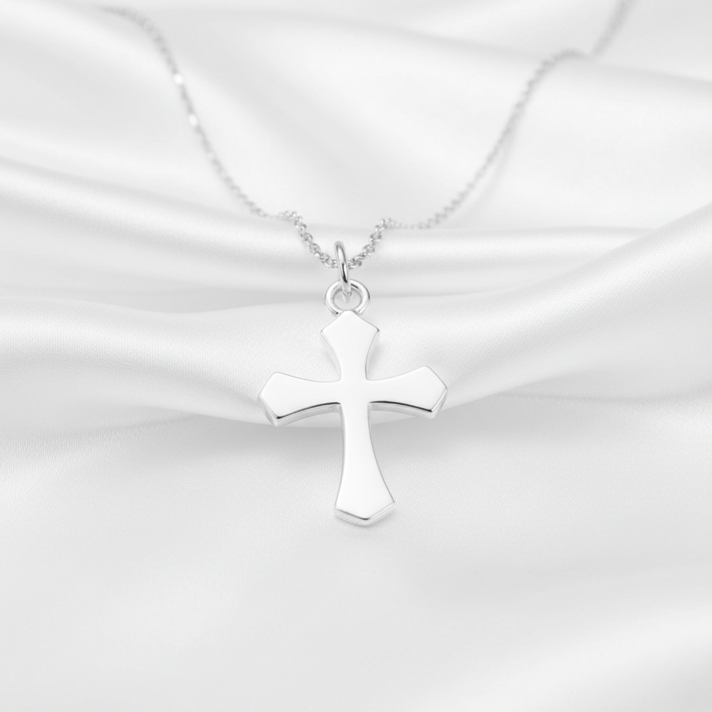 A polished silver cross pendant necklace on a delicate silver chain, displayed against a draped white satin fabric background.