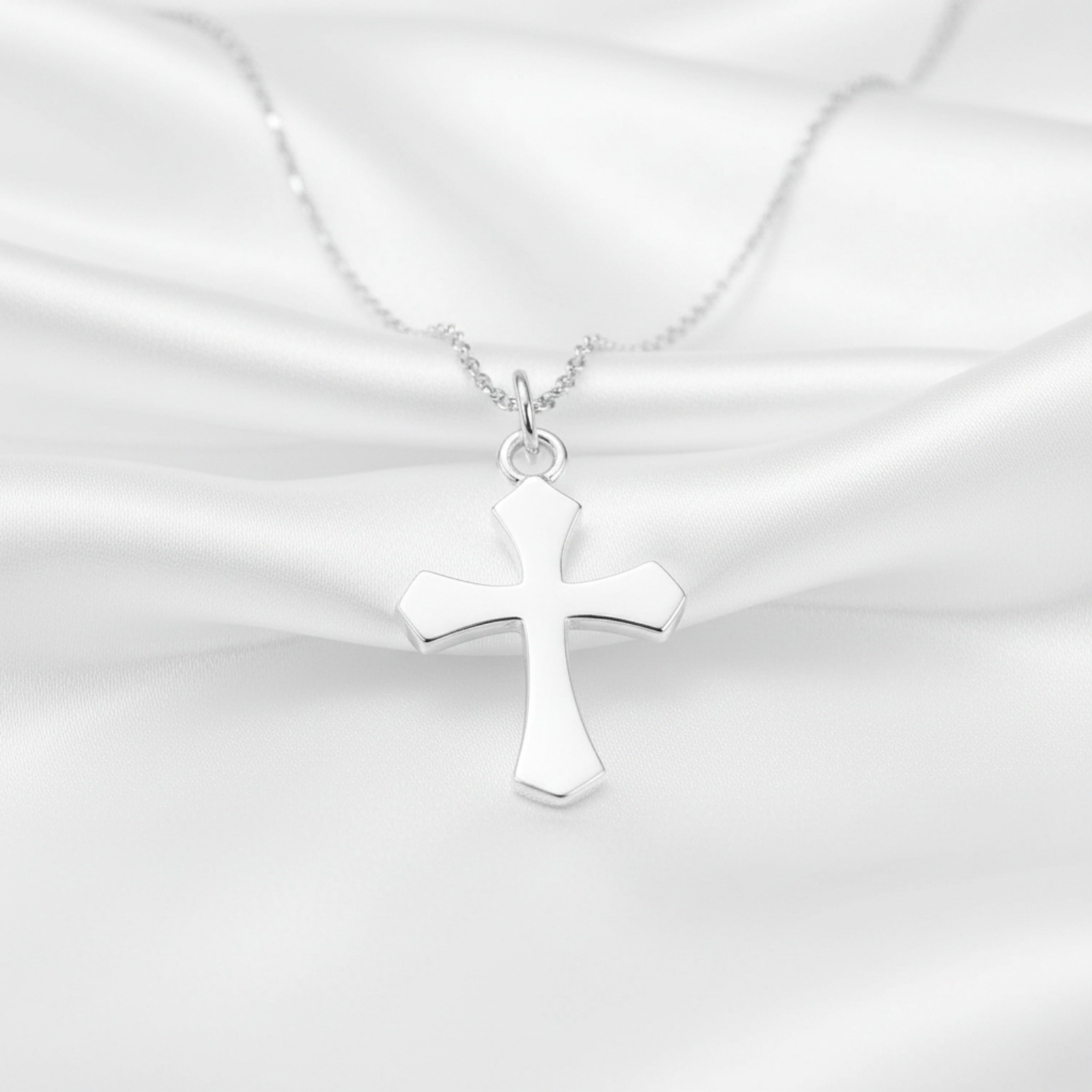 A polished silver cross pendant necklace on a delicate silver chain, displayed against a draped white satin fabric background.