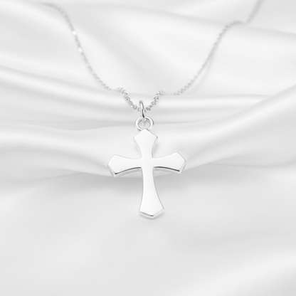 A polished silver cross pendant necklace on a delicate silver chain, displayed against a draped white satin fabric background.