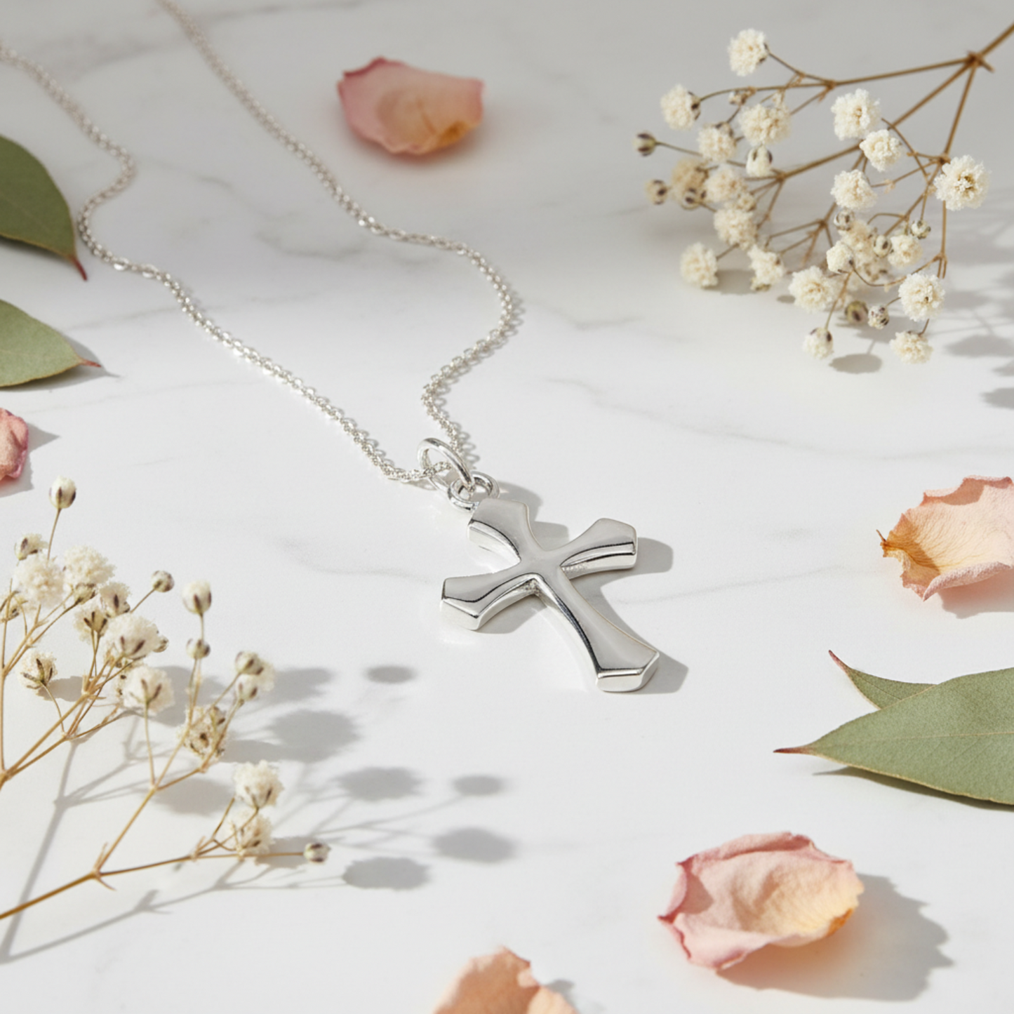A polished silver cross pendant necklace on a delicate silver chain, styled on a white marble surface with dried baby's breath flowers and pink rose petals.