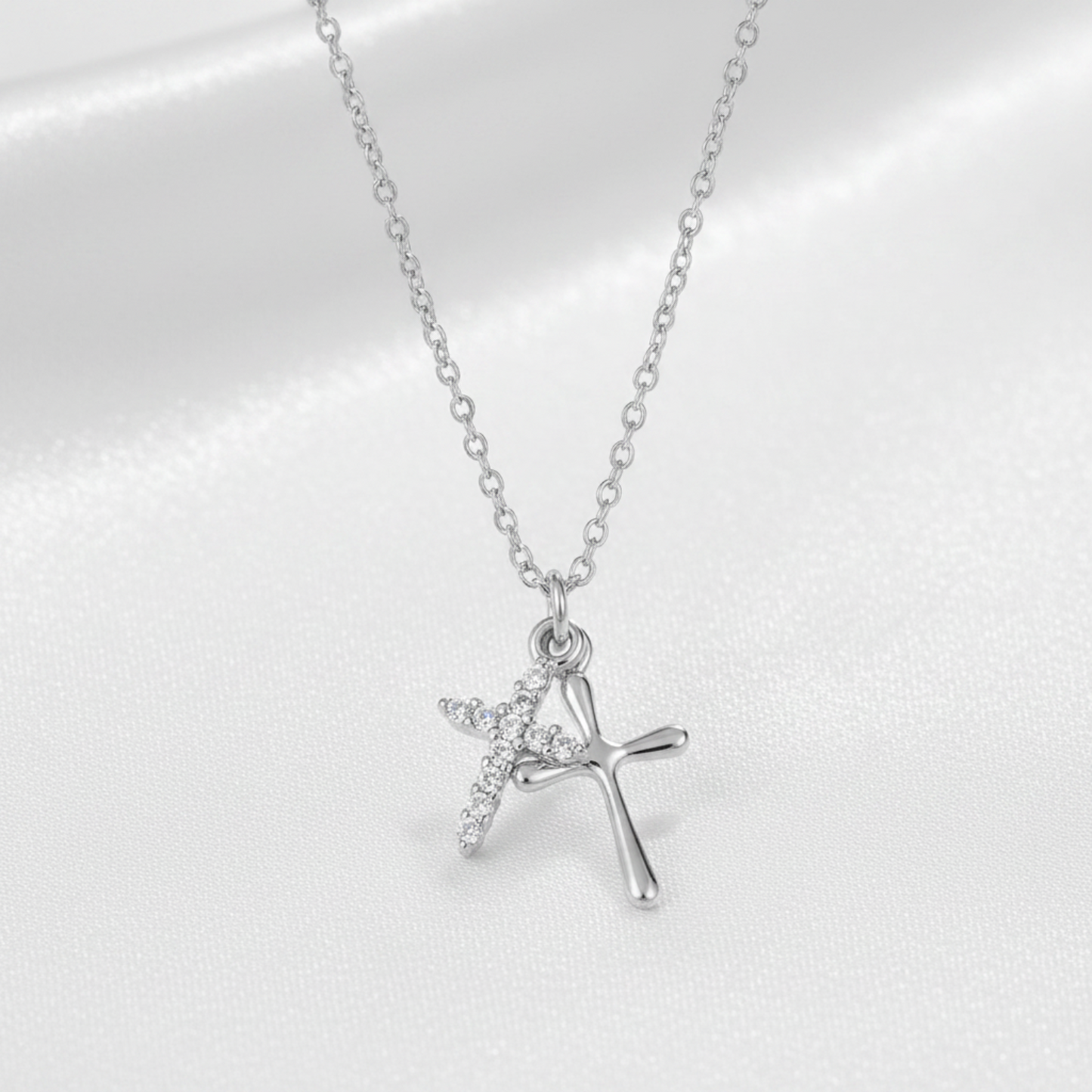 A silver cross pendant necklace with a small winged cross detail, displayed on a soft white satin fabric background.