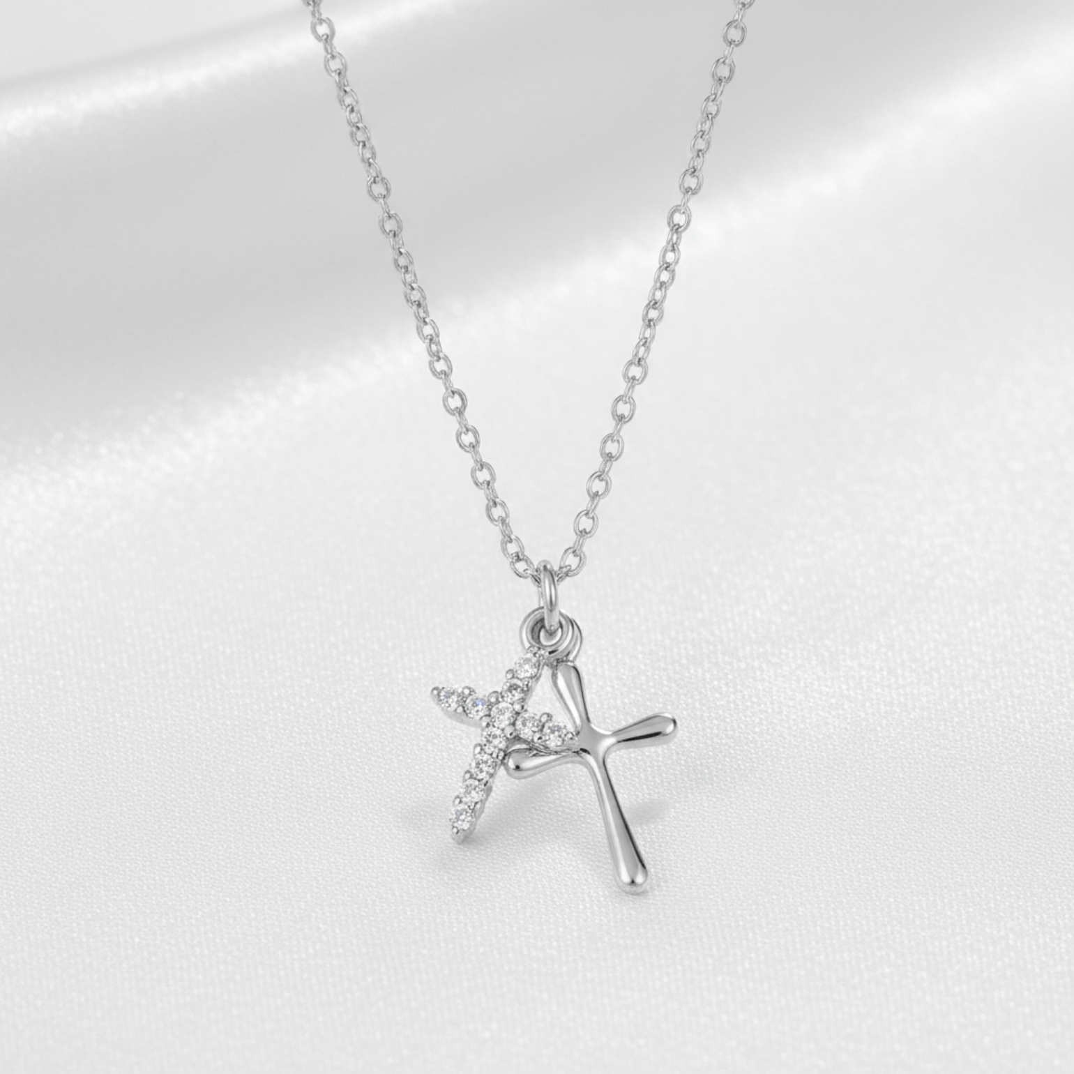 A silver cross pendant necklace with a small winged cross detail, displayed on a soft white satin fabric background.