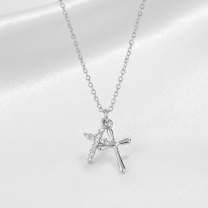 A silver cross pendant necklace with a small winged cross detail, displayed on a soft white satin fabric background.