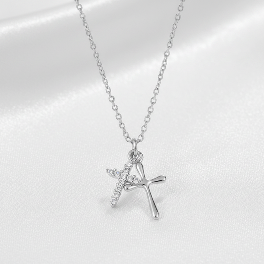 A silver cross pendant necklace with a small winged cross detail, displayed on a soft white satin fabric background.