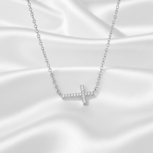A silver cross pendant necklace set with small clear stones, resting on a draped white satin fabric background.