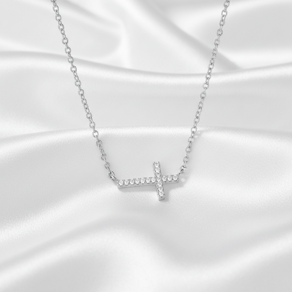 A silver cross pendant necklace set with small clear stones, resting on a draped white satin fabric background.