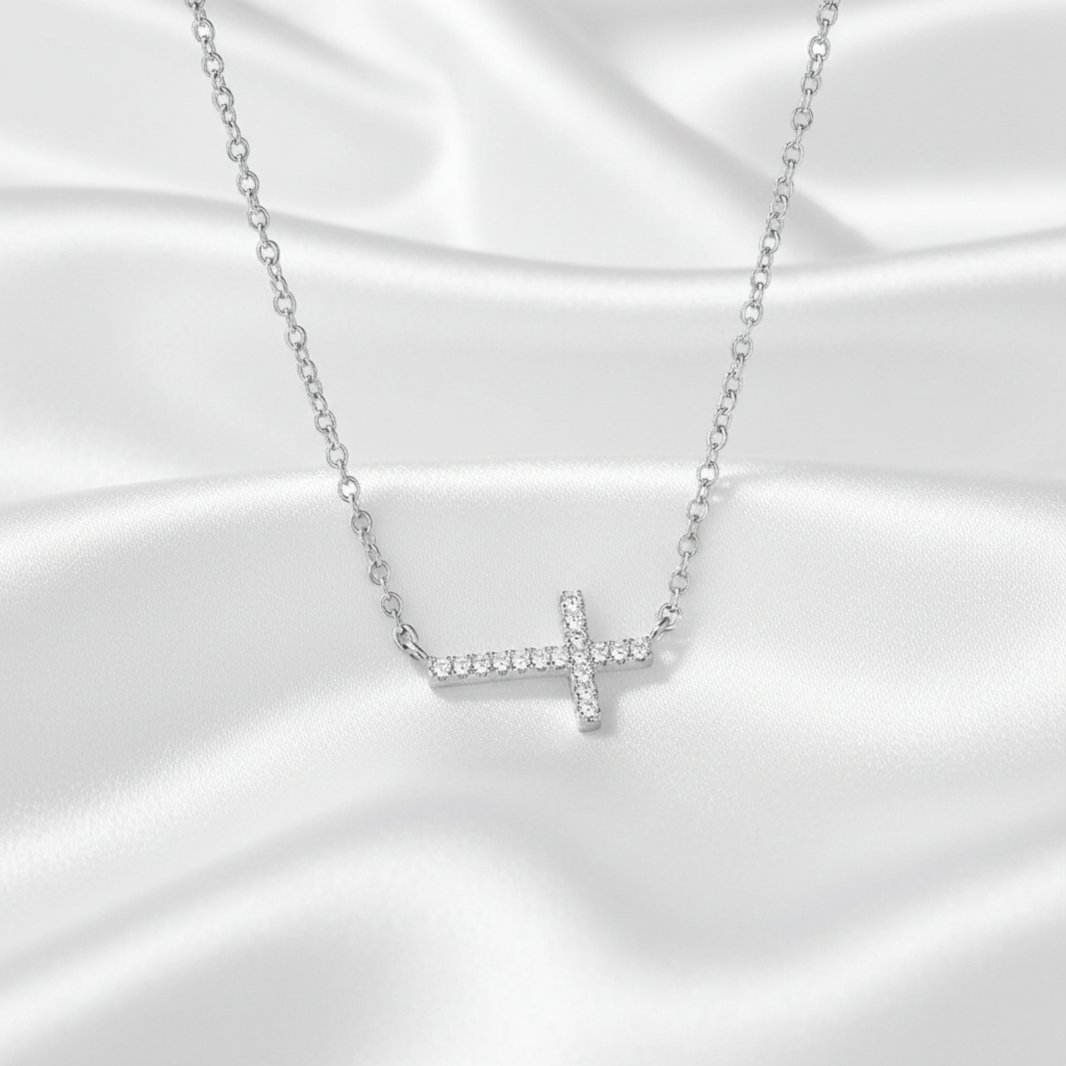 A silver cross pendant necklace set with small clear stones, resting on a draped white satin fabric background.