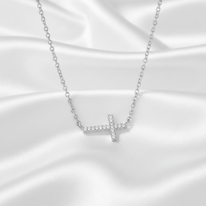 A silver cross pendant necklace set with small clear stones, resting on a draped white satin fabric background.