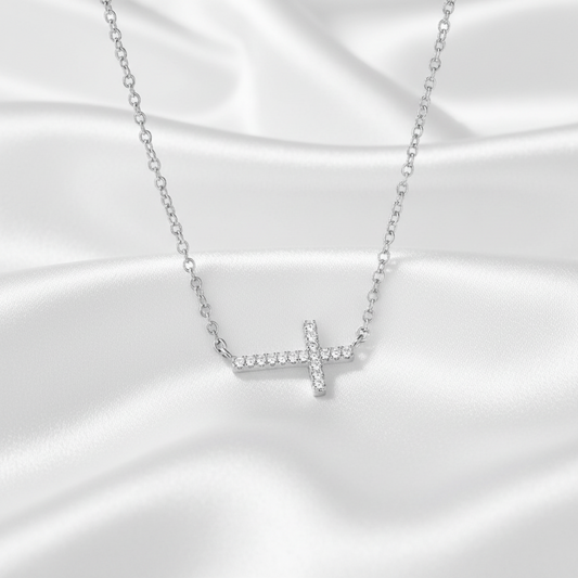 A silver cross pendant necklace set with small clear stones, resting on a draped white satin fabric background.