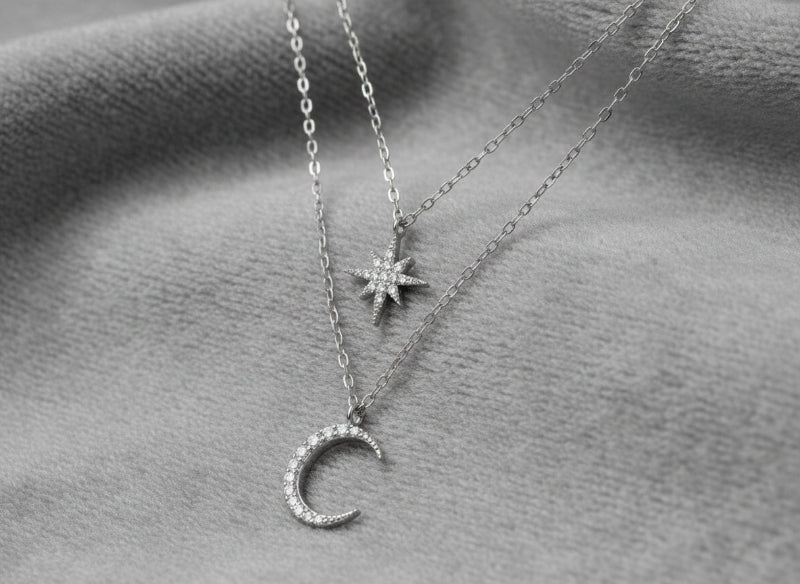 Two silver necklaces with star and moon pendants on a textured gray surface.
