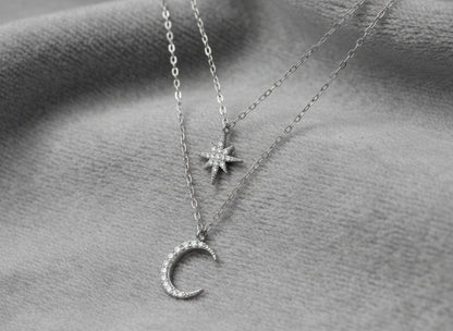 Two silver necklaces with star and moon pendants on a textured gray surface.