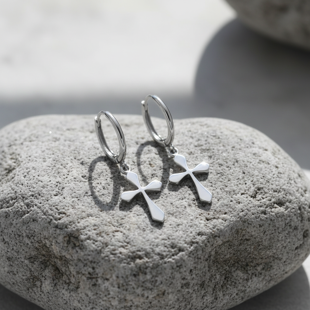 A pair of silver hoop earrings with small cross charms, resting on a textured gray stone with soft natural shadows.
