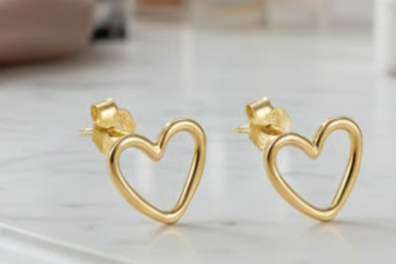 Gold heart-shaped earrings on a marble surface with a mirror and flowers in the background