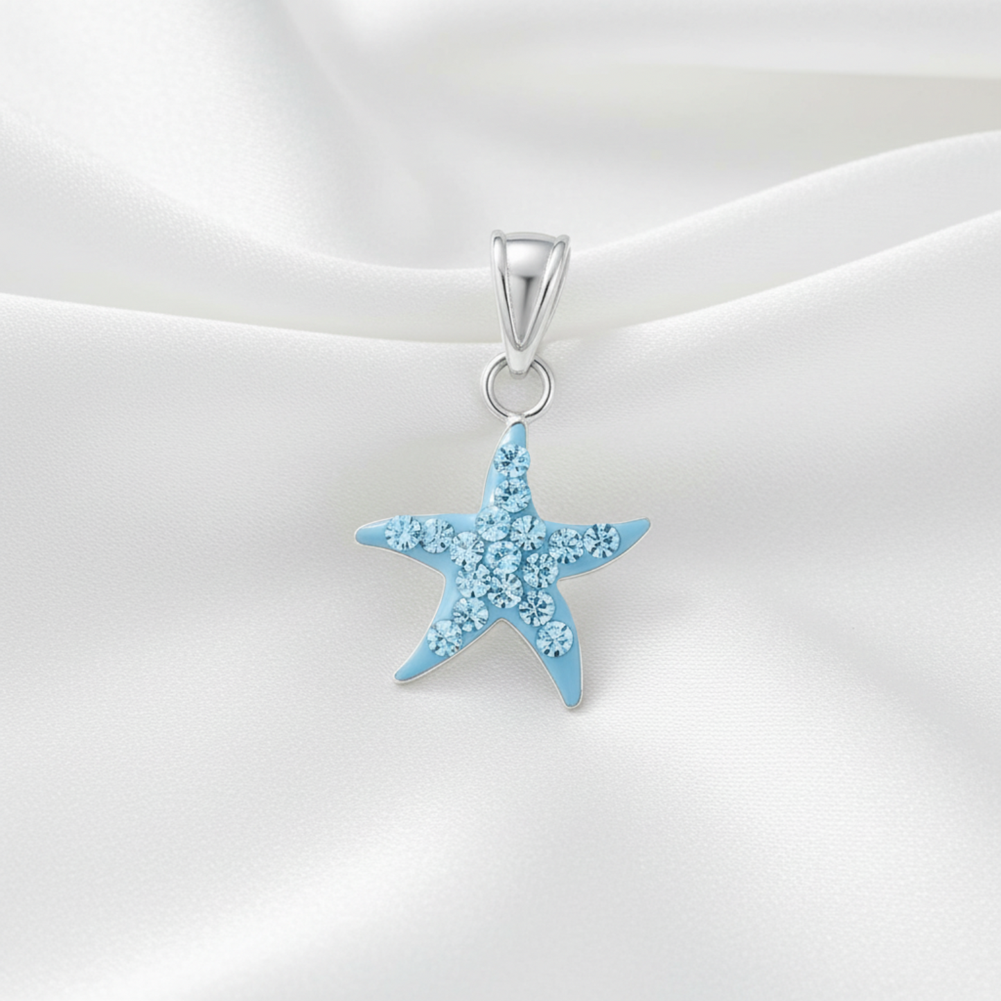 A light blue starfish-shaped pendant with sparkling clear rhinestones, suspended from a silver bail, displayed on a draped white satin fabric background.