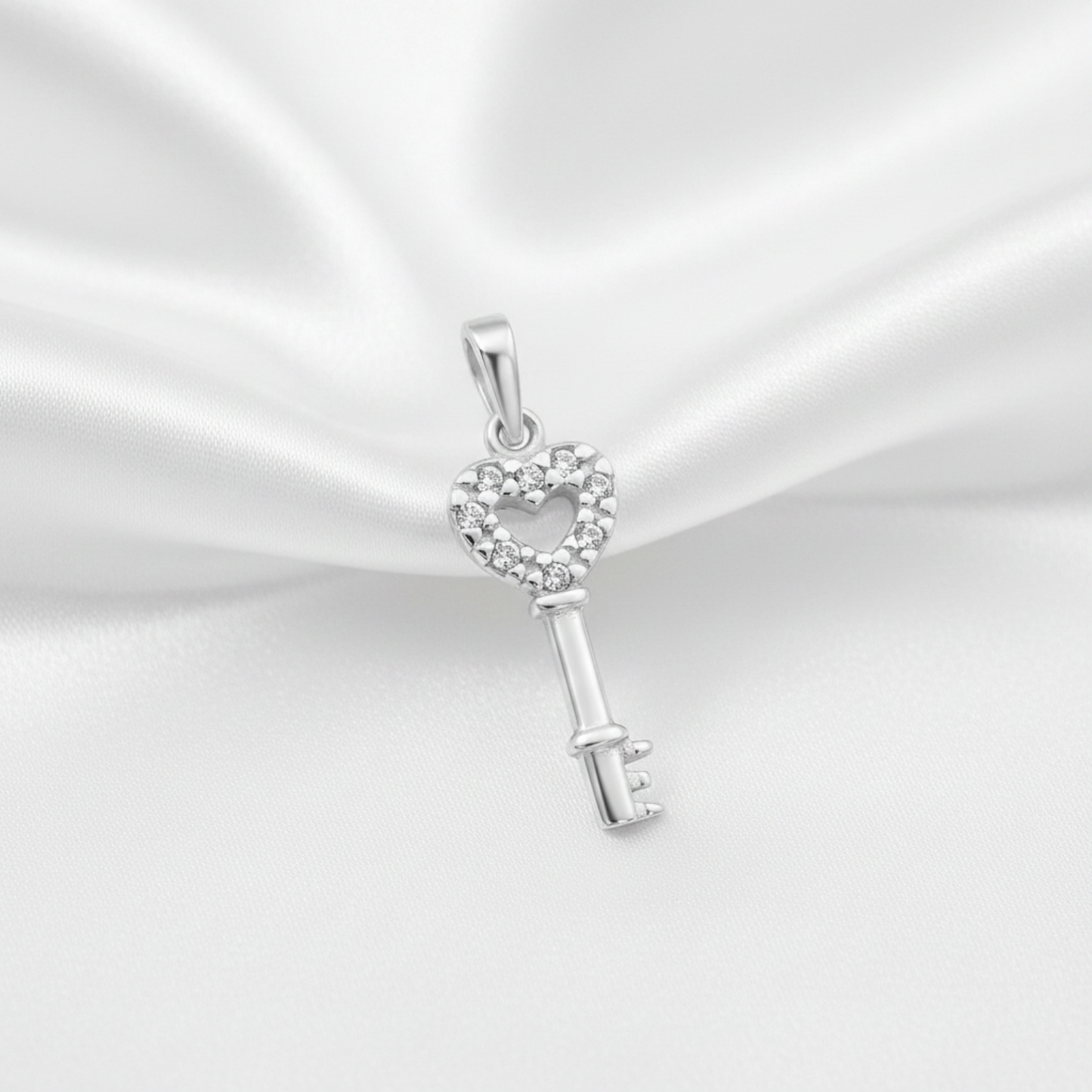 A silver key-shaped pendant with a heart-shaped head paved with small clear stones, displayed on a draped white satin fabric background.