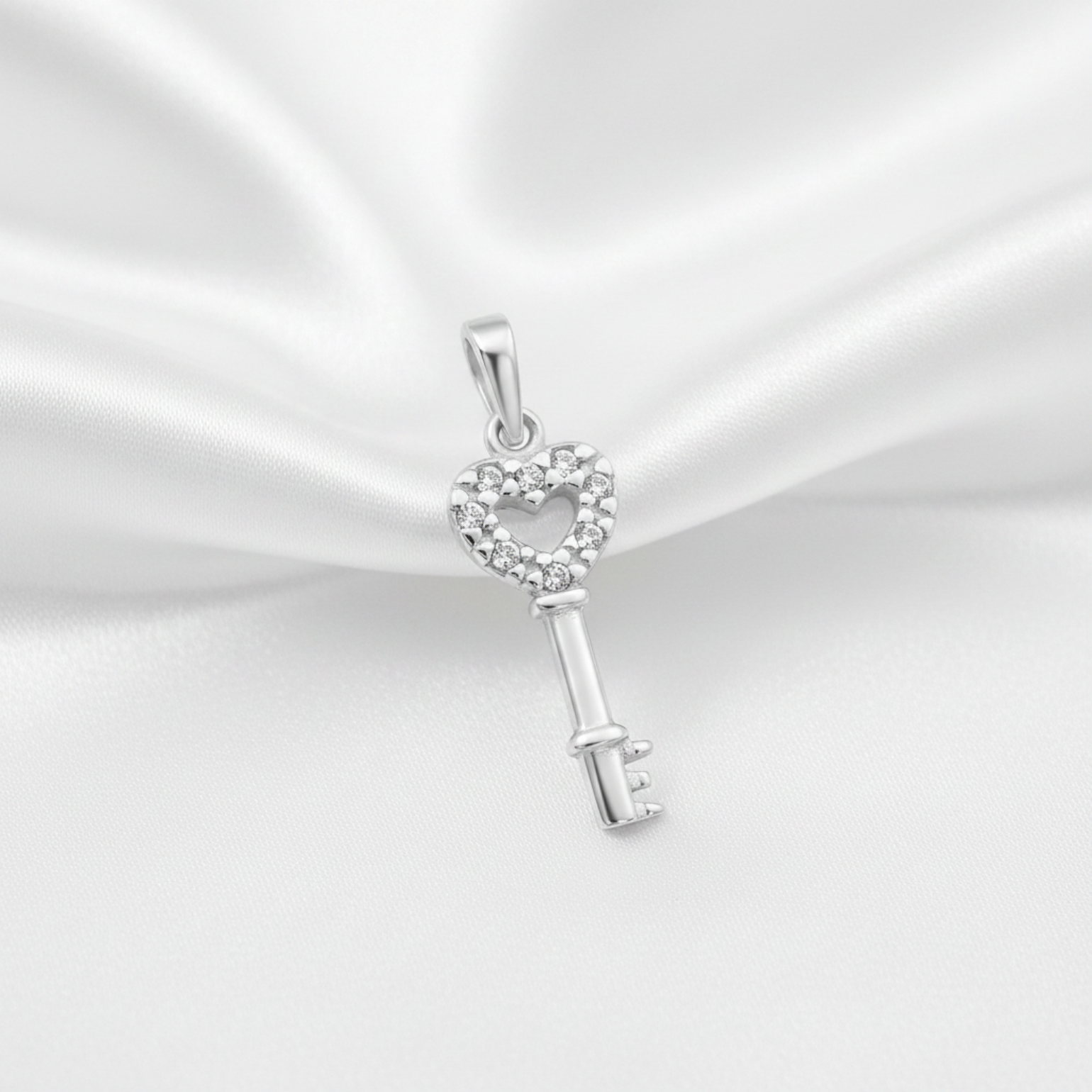 A silver key-shaped pendant with a heart-shaped head paved with small clear stones, displayed on a draped white satin fabric background.