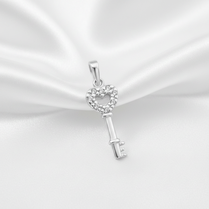 A silver key-shaped pendant with a heart-shaped head paved with small clear stones, displayed on a draped white satin fabric background.