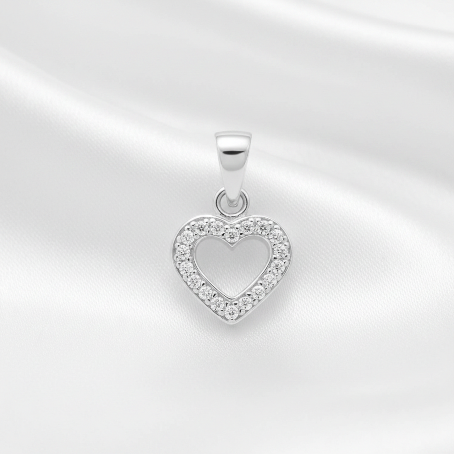 A silver heart-shaped pendant with a border of small clear stones, displayed on a white satin fabric background.