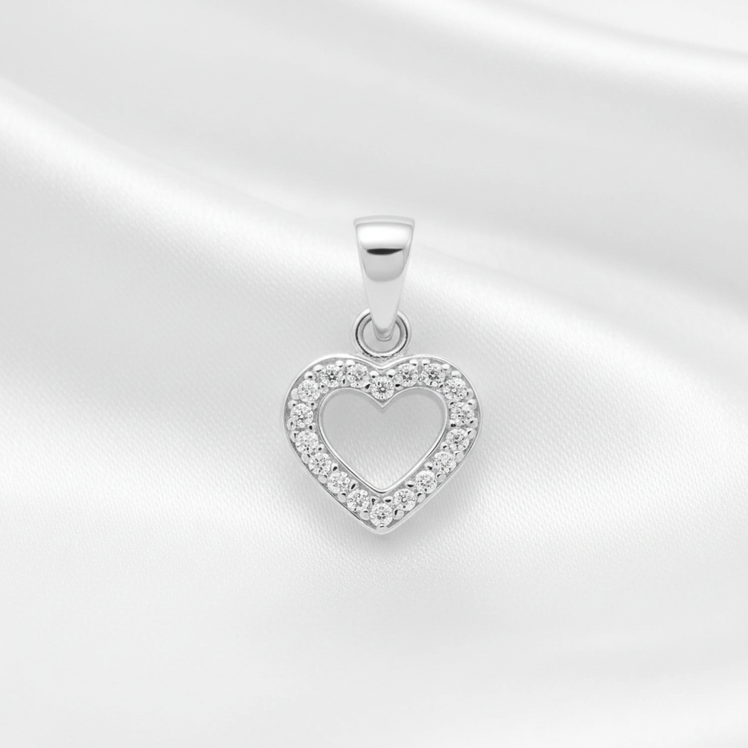A silver heart-shaped pendant with a border of small clear stones, displayed on a white satin fabric background.