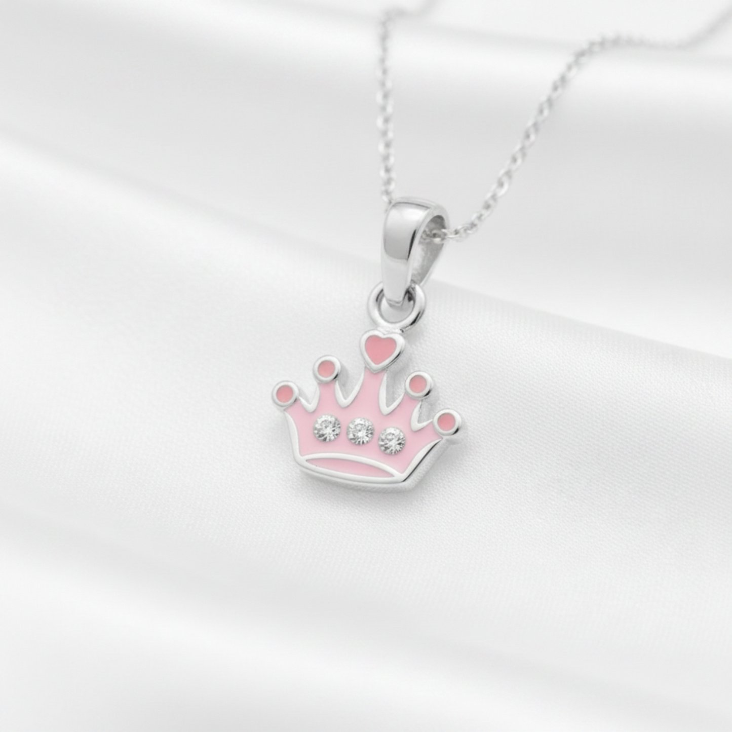 A silver crown pendant necklace with pink enamel details and small clear rhinestones, displayed on a white satin fabric background.