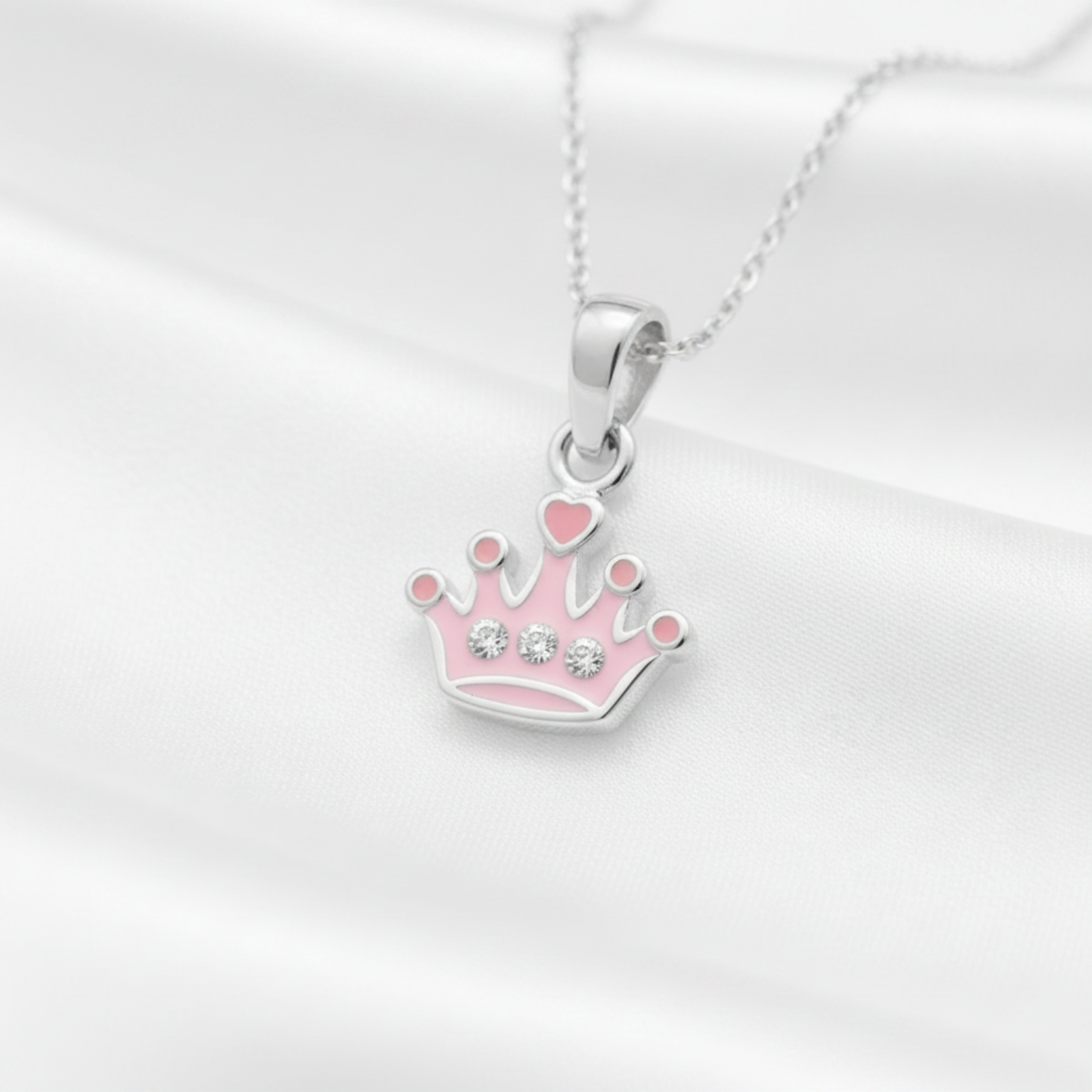 A silver crown pendant necklace with pink enamel details and small clear rhinestones, displayed on a white satin fabric background.
