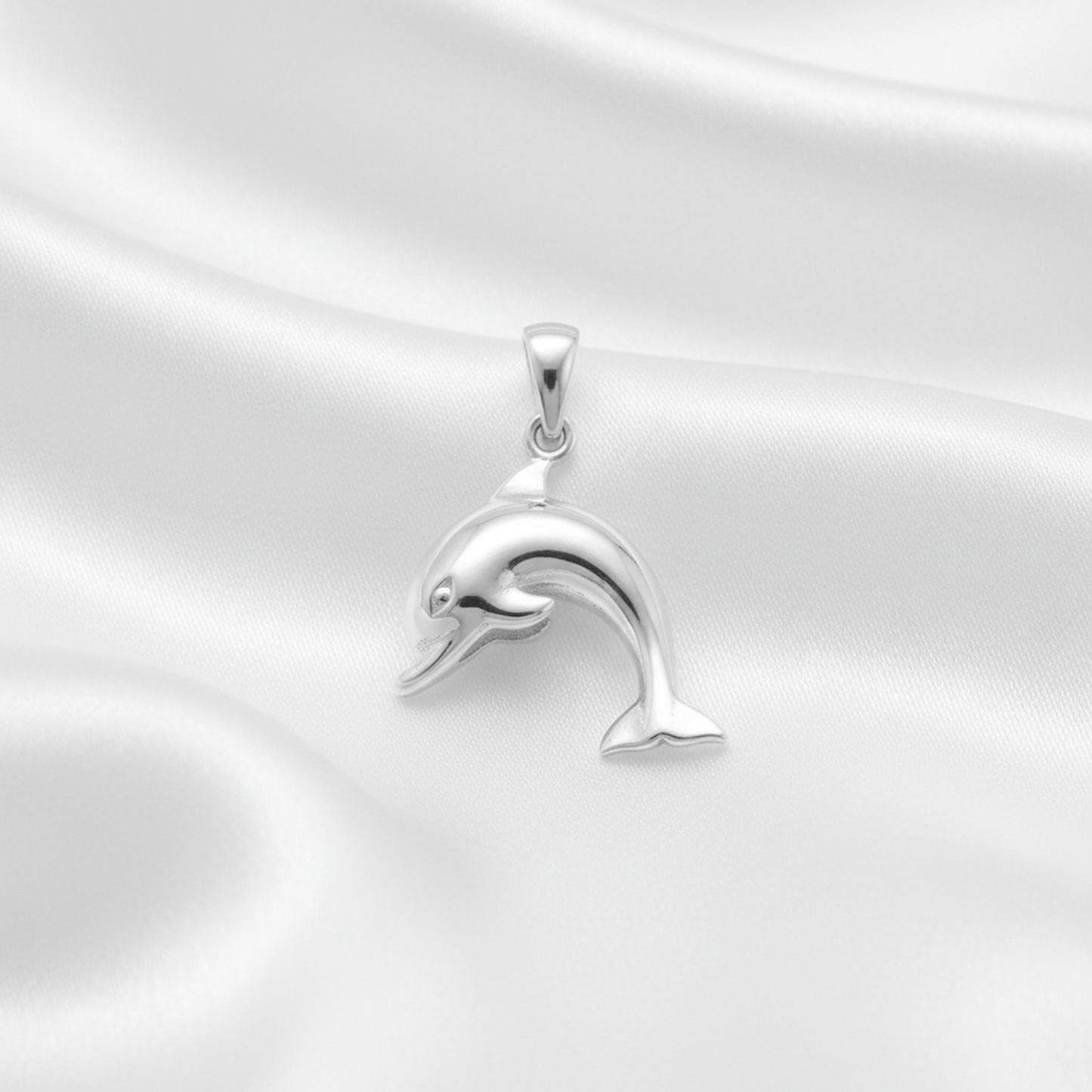 A polished silver dolphin pendant with a smooth, reflective finish, resting on a draped white satin fabric background.