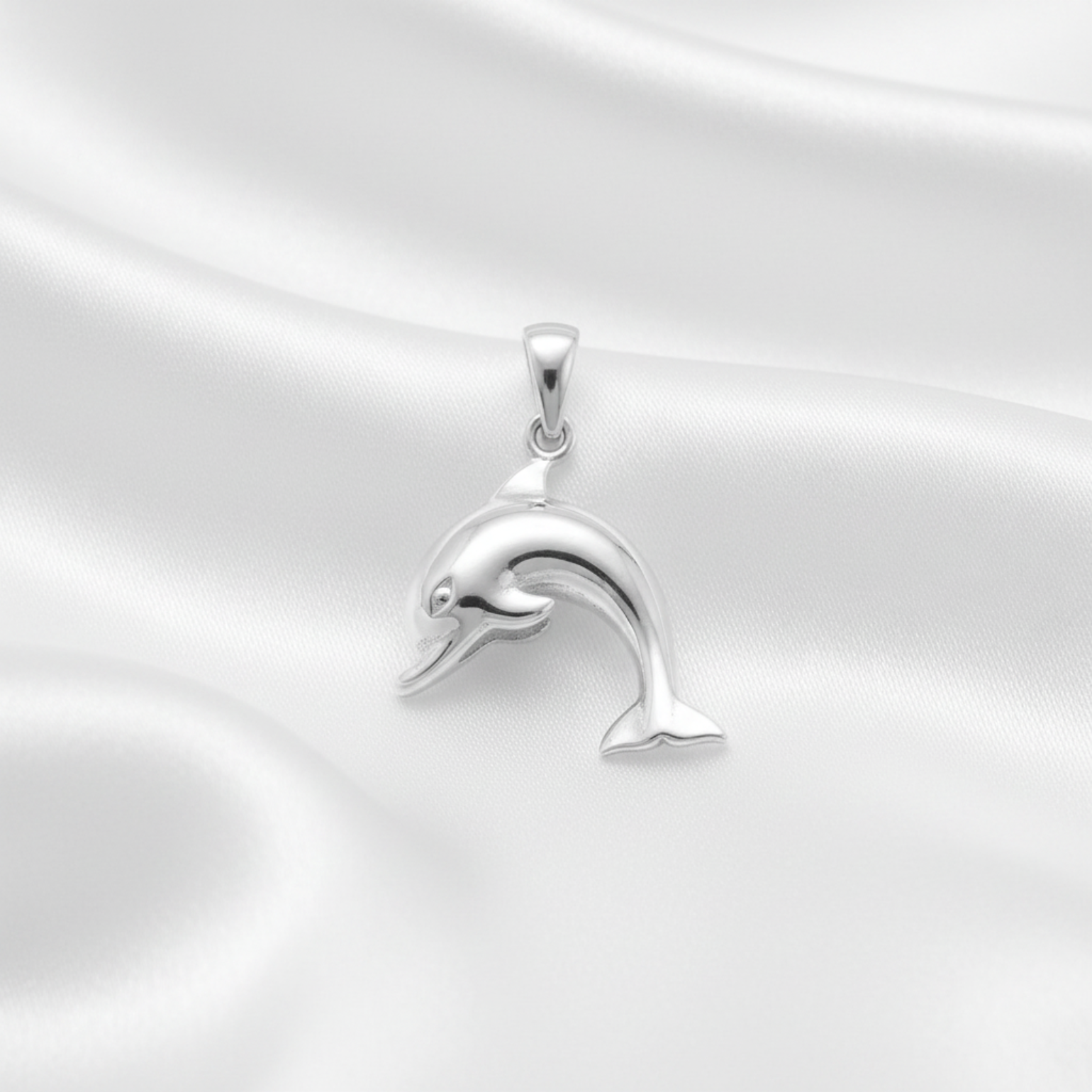 A polished silver dolphin pendant with a smooth, reflective finish, resting on a draped white satin fabric background.