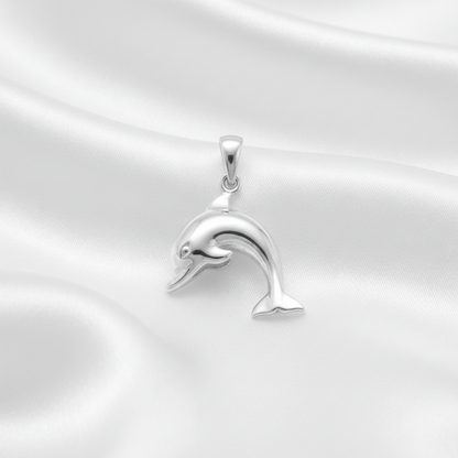 A polished silver dolphin pendant with a smooth, reflective finish, resting on a draped white satin fabric background.