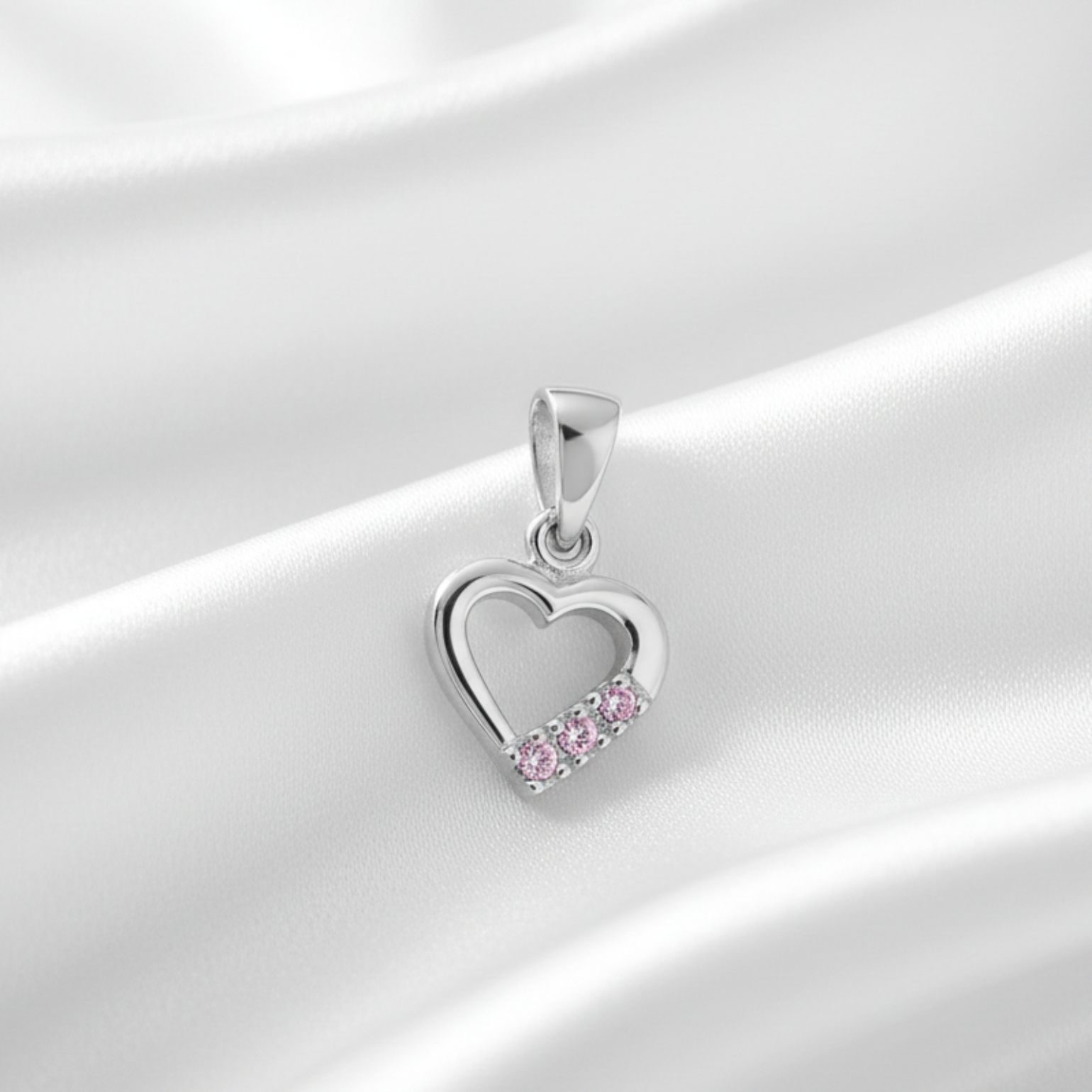A silver heart-shaped pendant with a row of small pink gemstones along the bottom curve, displayed on draped white satin fabric.