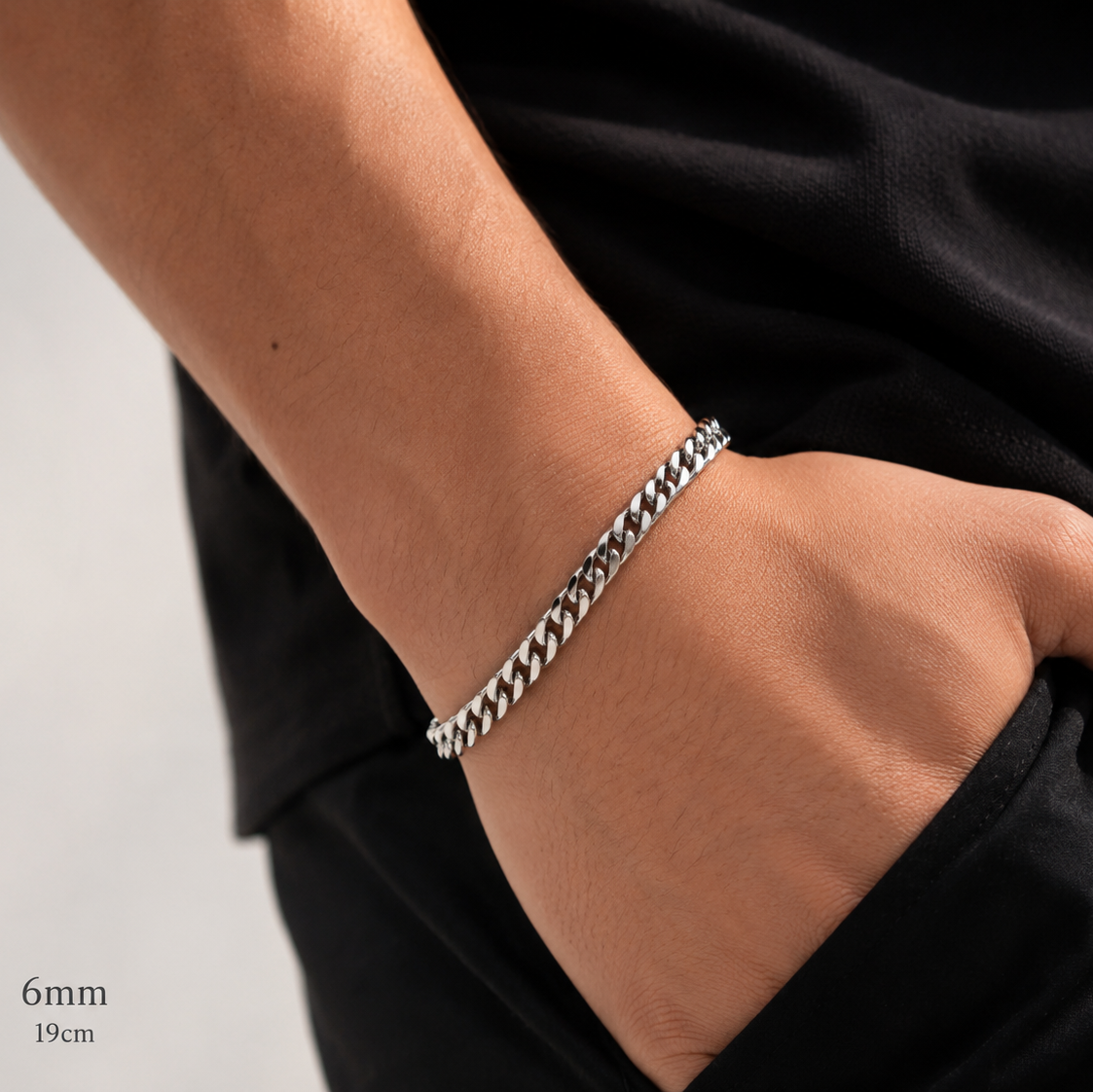 A close-up of a person's wrist wearing a silver-toned curb chain bracelet, with their hand resting in a black pocket. 
