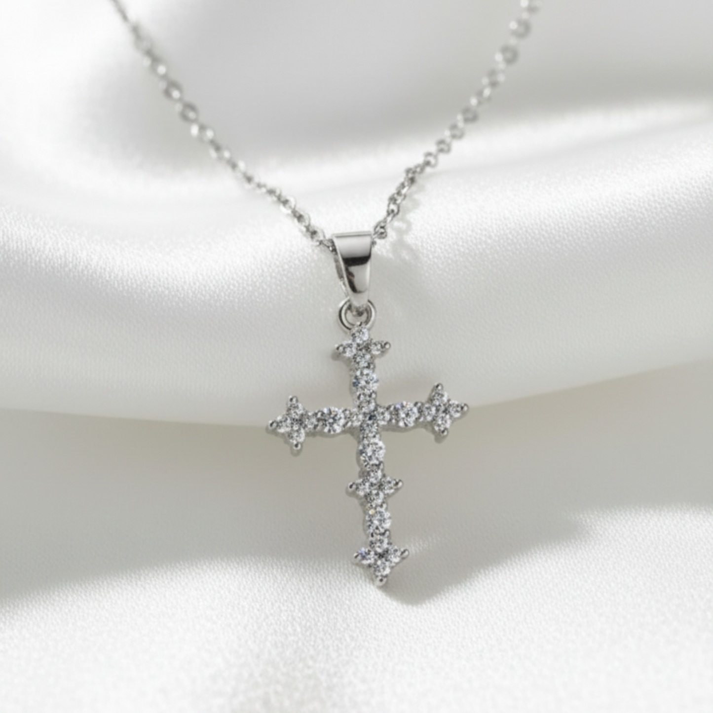 A silver cross pendant necklace adorned with small clear stones, displayed on a draped white satin fabric background.