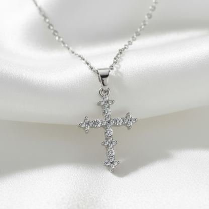 A silver cross pendant necklace adorned with small clear stones, displayed on a draped white satin fabric background.