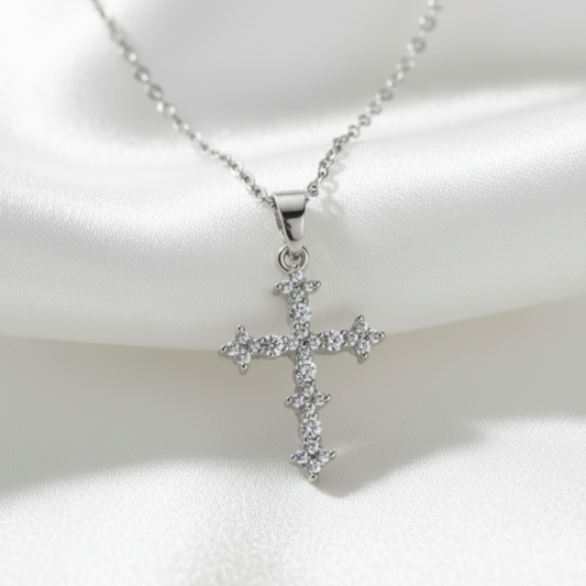 A silver cross pendant necklace adorned with small clear stones, displayed on a draped white satin fabric background.