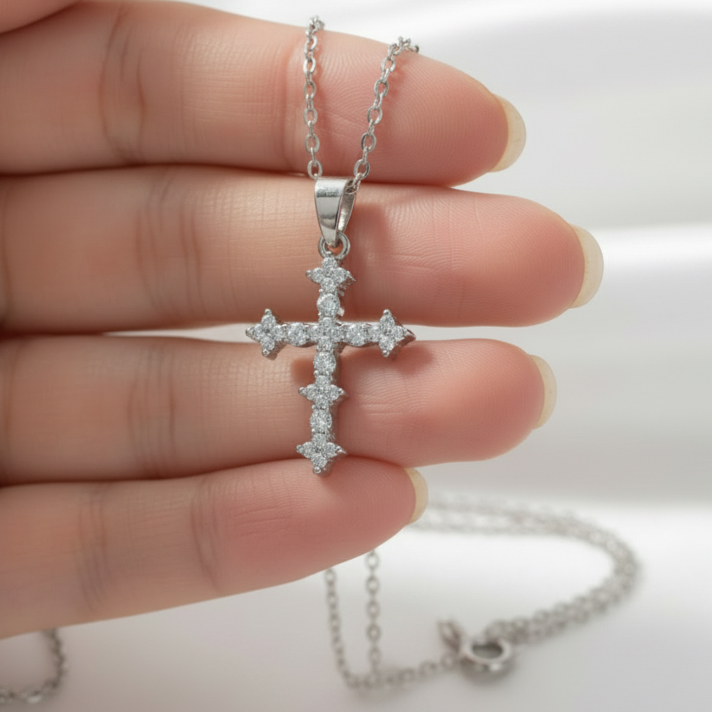 A silver cross pendant necklace with small clear stones, held in a person's hand against a soft white background.