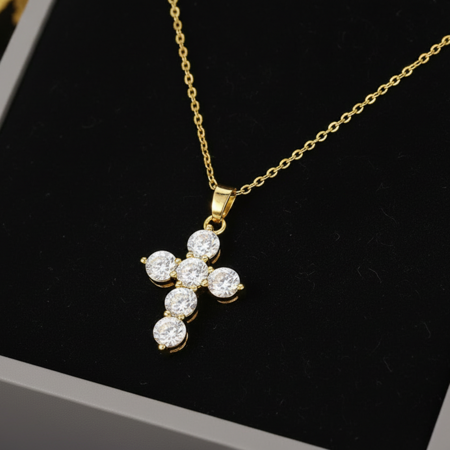 A gold cross pendant necklace with five clear round-cut stones, displayed on a black velvet insert inside a gray jewelry box.
