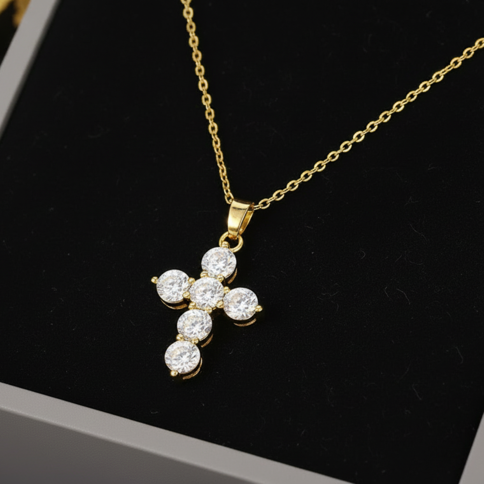 A gold cross pendant necklace with five clear round-cut stones, displayed on a black velvet insert inside a gray jewelry box.