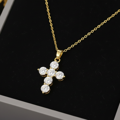 A gold cross pendant necklace with five clear round-cut stones, displayed on a black velvet insert inside a gray jewelry box.