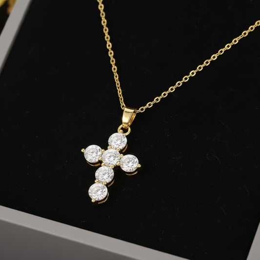 A gold cross pendant necklace with five clear round-cut stones, displayed on a black velvet insert inside a gray jewelry box.