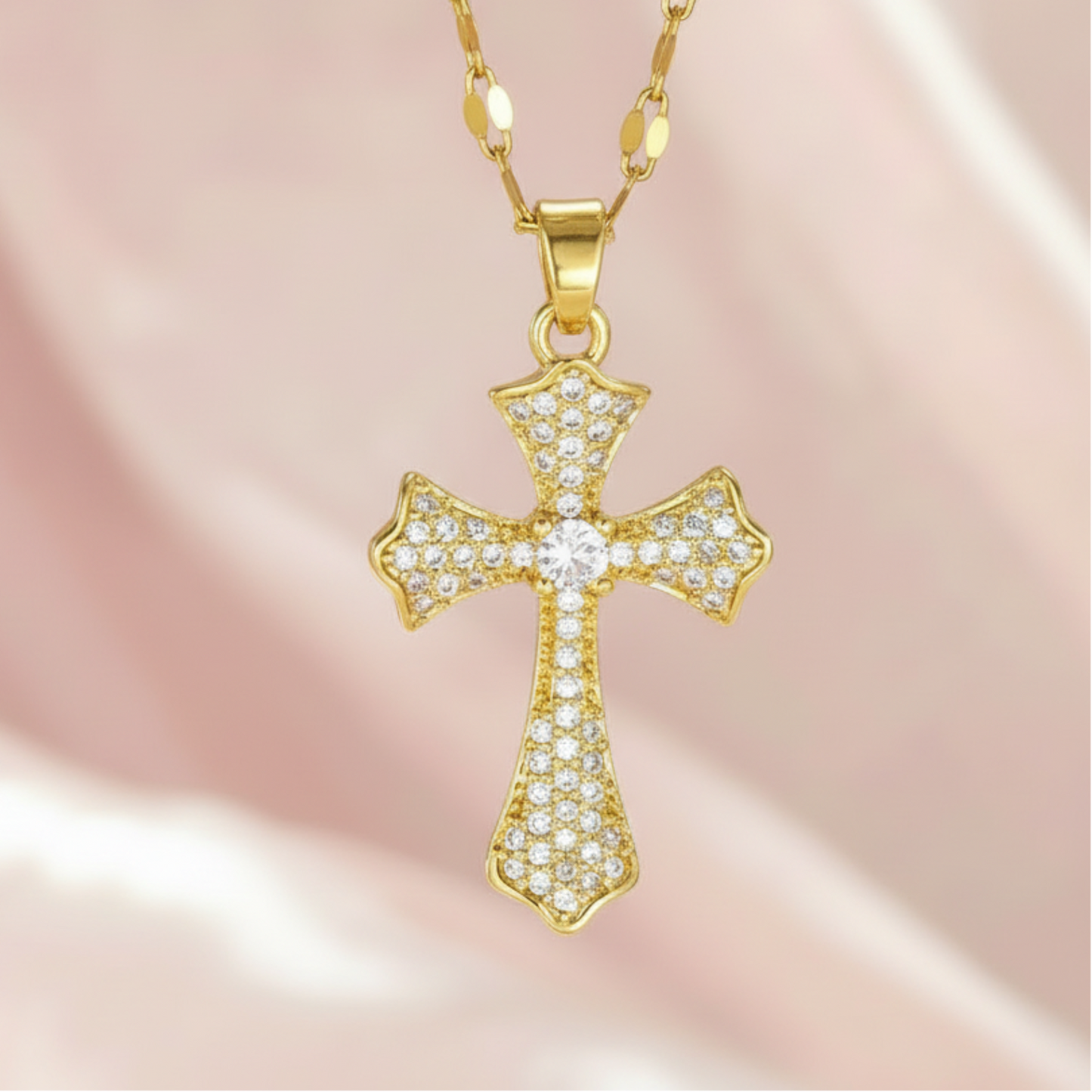 A gold cross pendant necklace paved with small clear stones, hanging from a gold chain against a soft pink satin background.